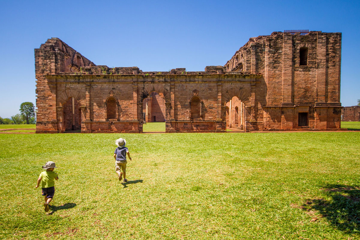 two young boys run across a field towards the ruins of a Jesuit mission in Paraguay