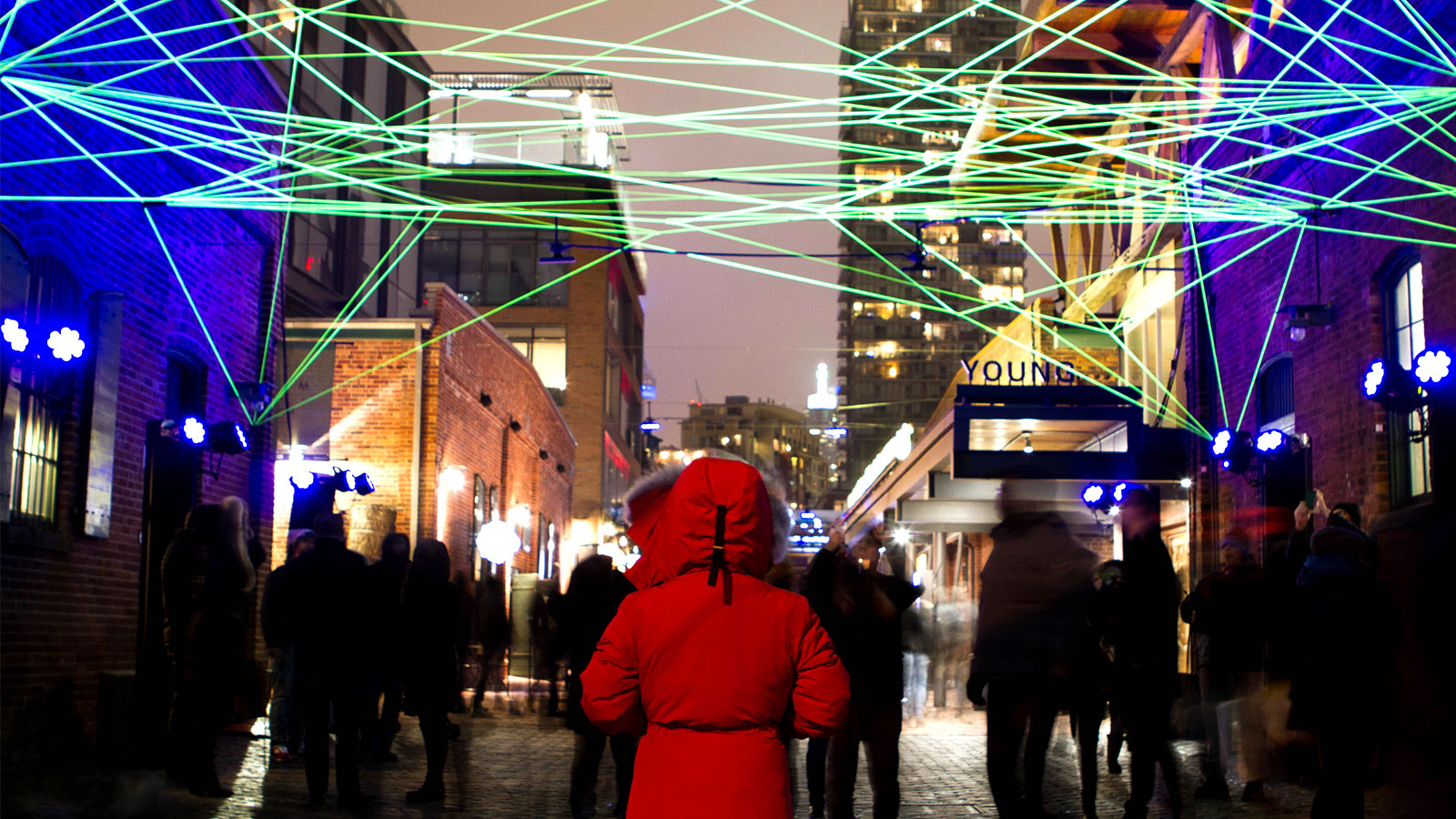 A woman in a red Canada Goose Jacket looks down an alley under a web of green light at the Toronto LightFest in the Distillery District