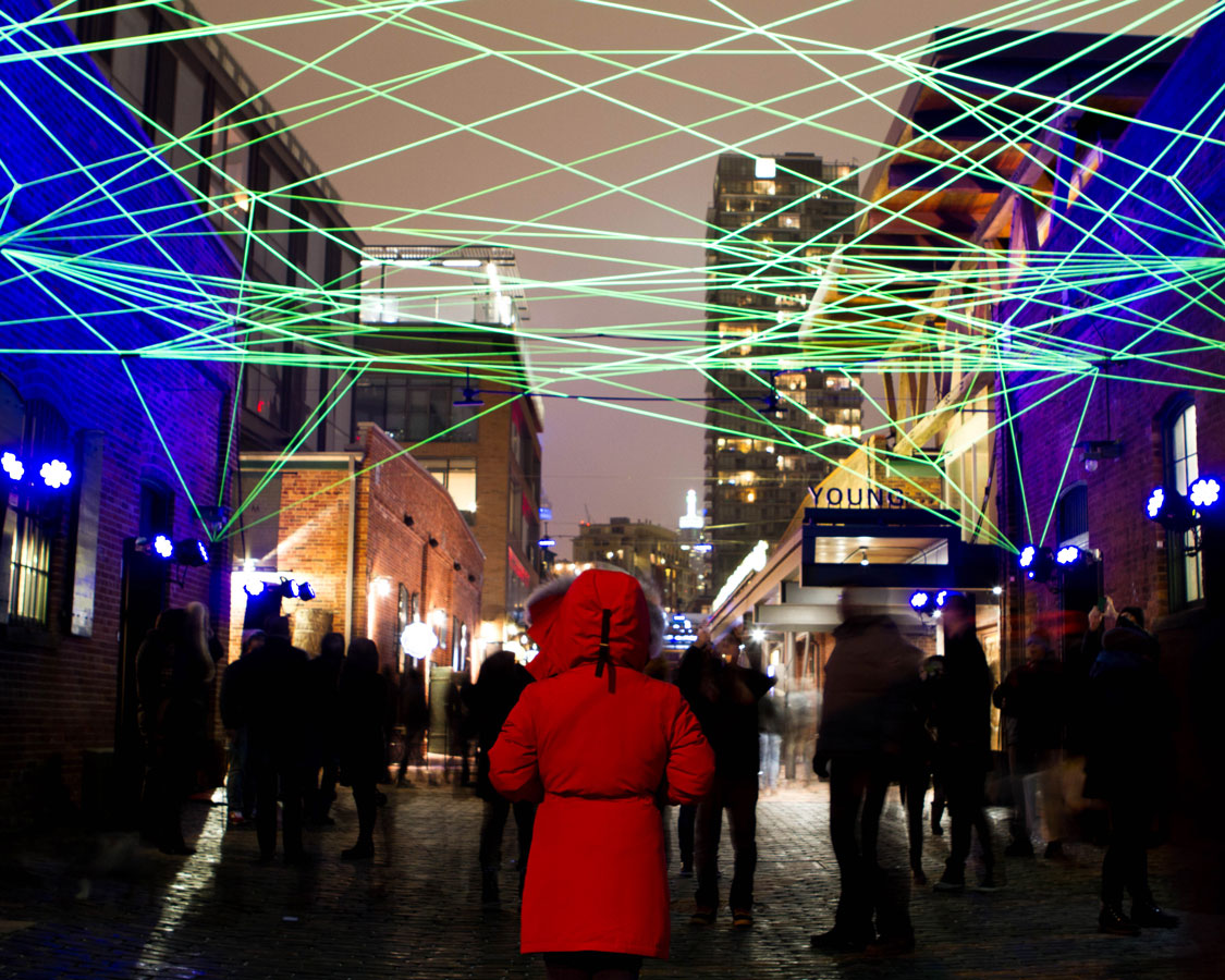 A woman in a red Canada Goose Jacket looks down an alley under a web of green light at the Toronto LightFest in the Distillery District