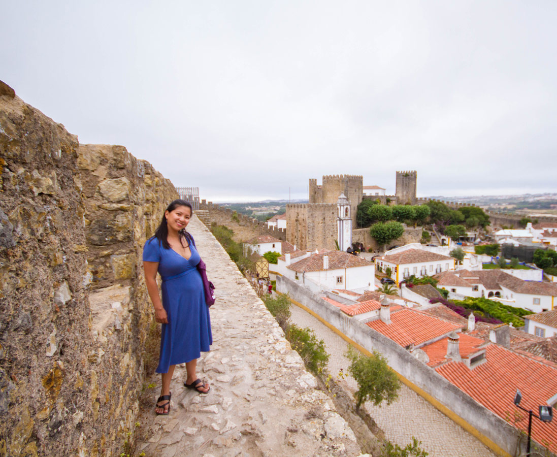 A woman in a blue dress stands on the inside walls of the city of Obidos, Portugal while the castle stands in the background