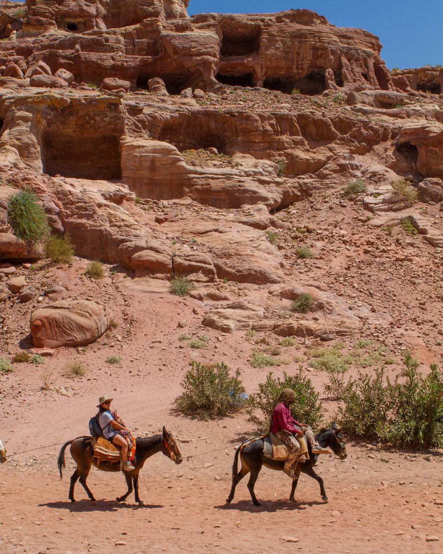 A Bedhouin guide and a woman on horseback ride past cave houses in Petra Jordan