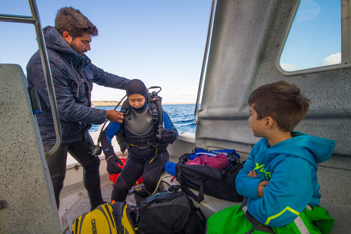 A young boy watches his mother put on SCUBA gear with the help of a dive master as she prepares for Scuba diving with sea lions in Punta Loma