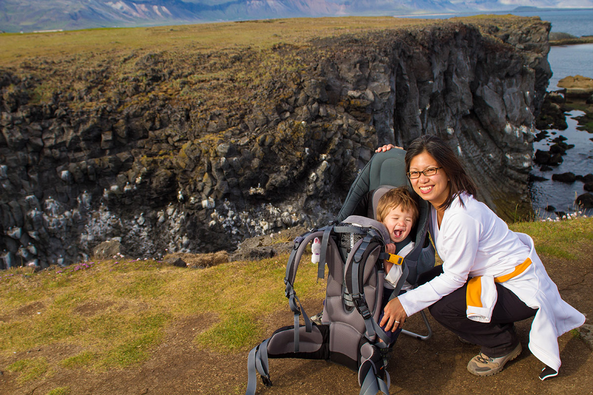 A mother smiles kneeling next to a young boy in a kid-carrier on a hike in Arnarpstapi Iceland