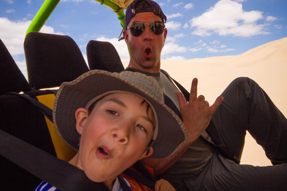 A father and son celebrate while riding in a dune buggy through the desert as they prepare to go sandboarding with kids in Peru in the desert near Huacachina Peru