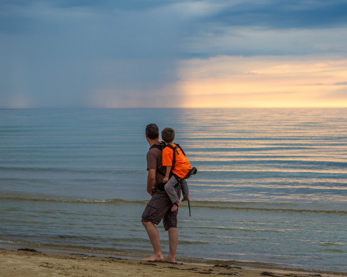 A father carries a child along a beach. The child sitting high on the Freeloader Child Carrier as they pass by a sunset and rain clouds