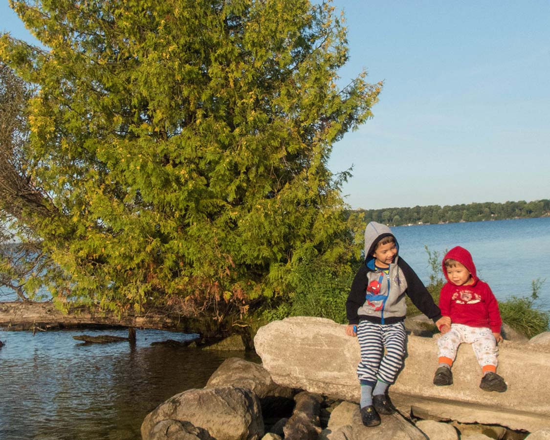 A young boy and a toddler hold hands while sitting on rocks near a tree and lake - Mara Provincial Park