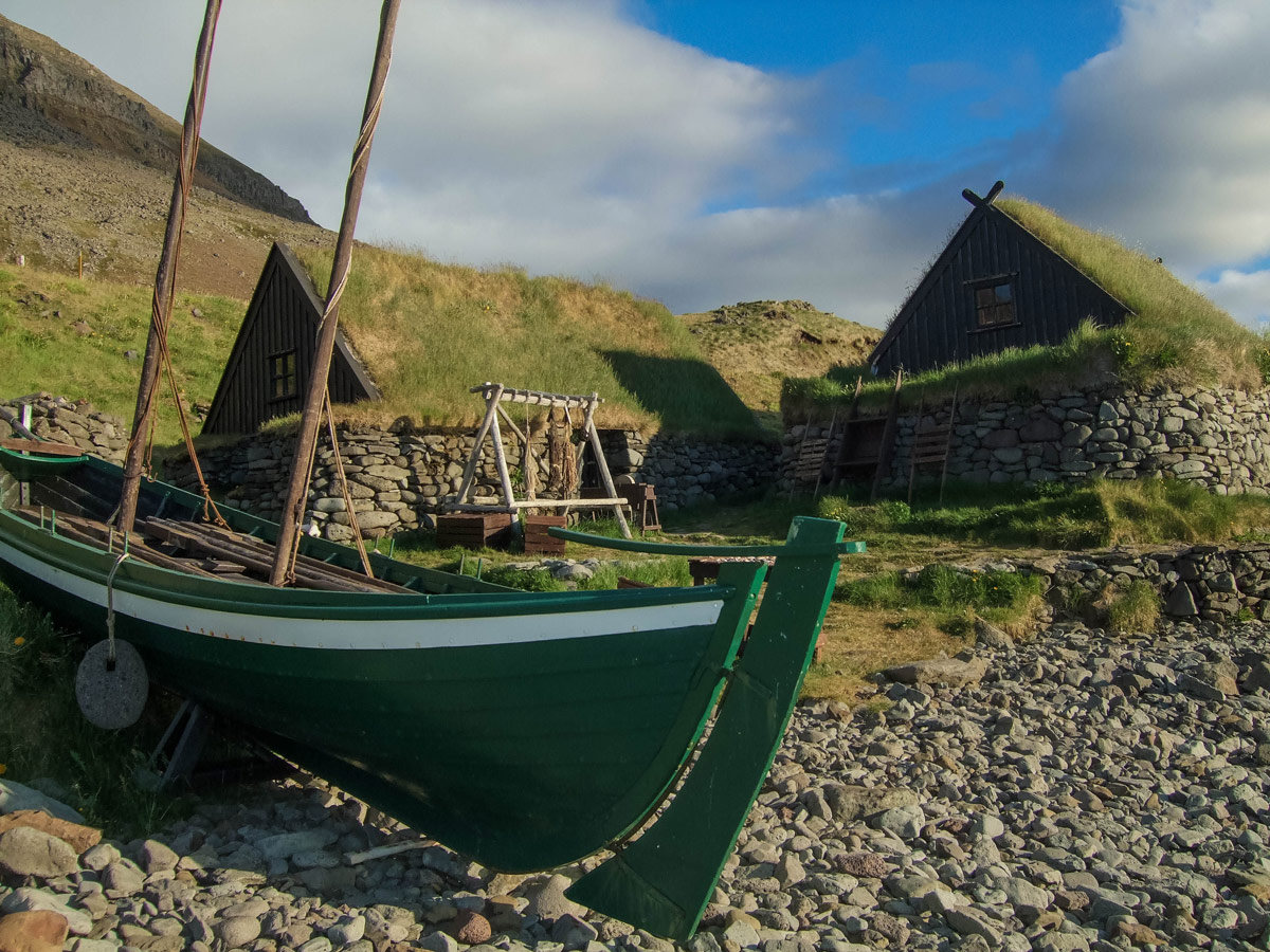 A wooden fishing boat sits in front of grass covered fishing huts on the coast of Iceland's westfjords