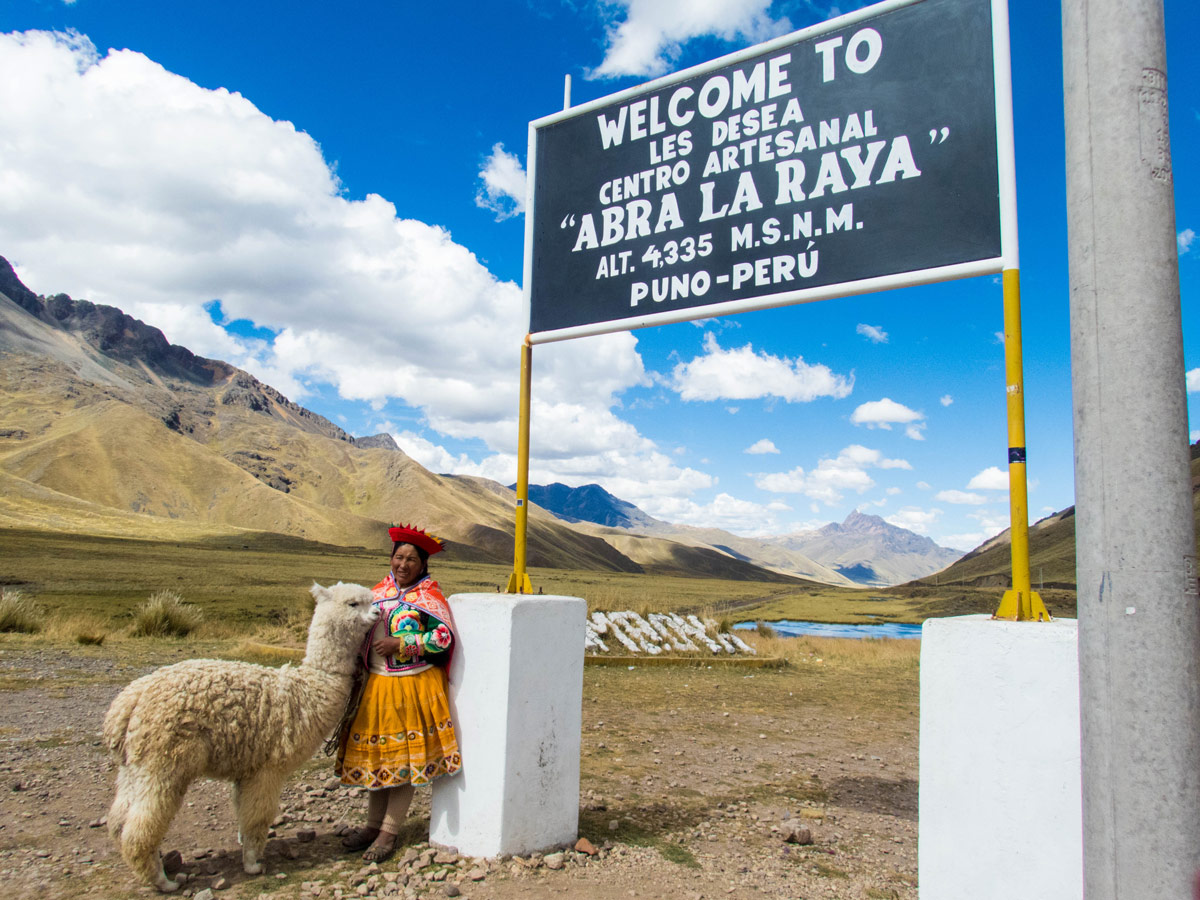 A Peruvian Quechua woman stands with a llama next to a sign in La Raya Peru on the Cusco to Puno bus tour