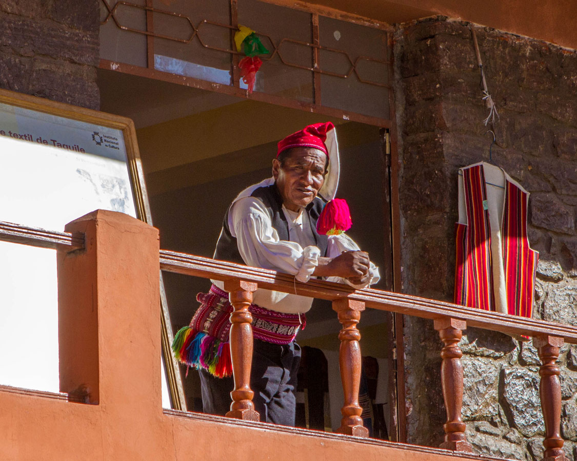 A Taquileano man wearing a black vest and colourful hat and belt stands outside a garment shop on Isla Taquile on Lake Titicaca Peru