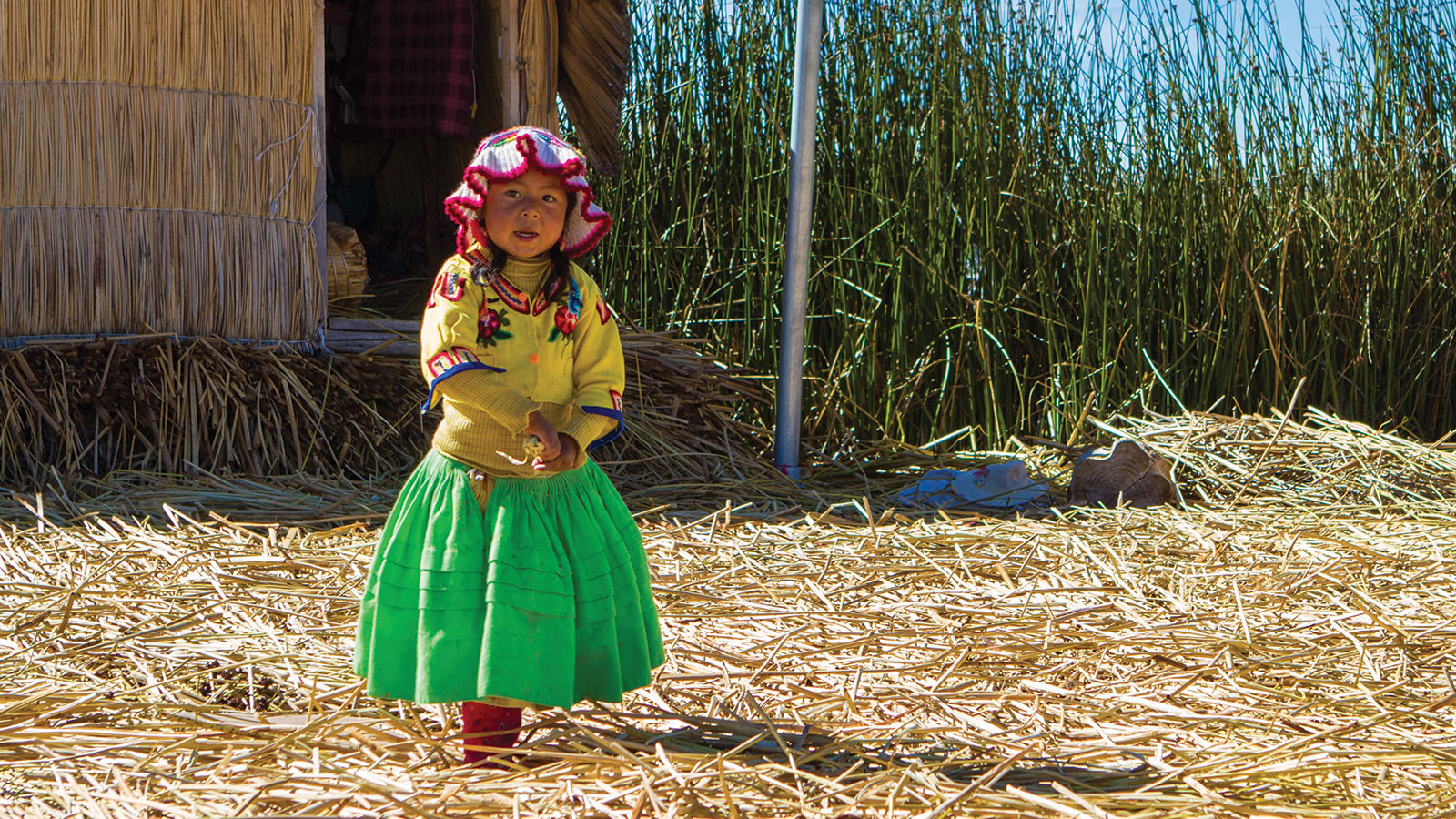 A young Uru girl in traditional clothing walks on a reed island in Isla de los Uros on Lake Titicaca with kids