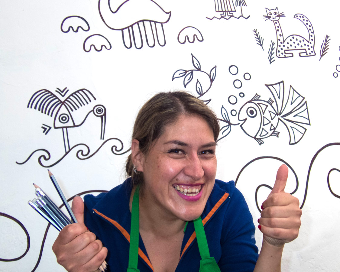 A Peruvian artist smiles and gives two thumbs up in front of a wall of inca style sketches