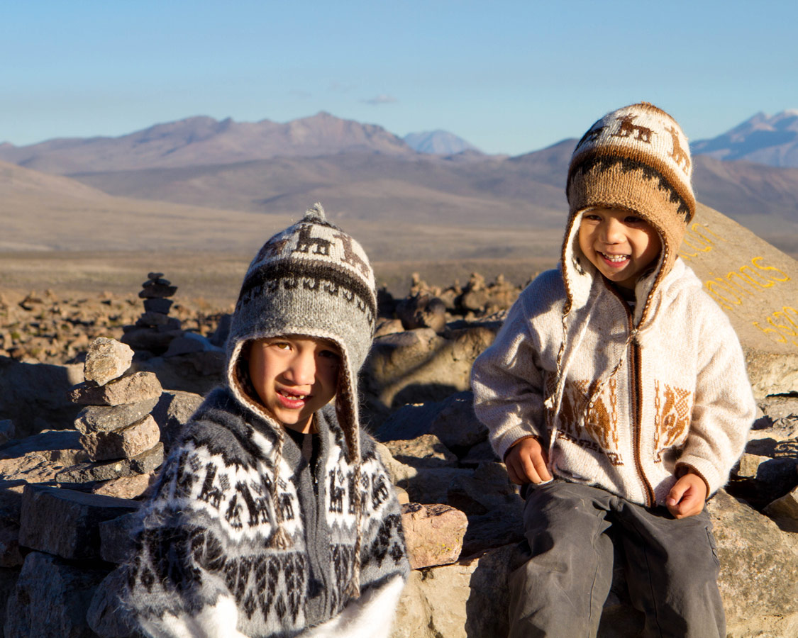 Two young boys wear alpaca hats and sweaters smile among piledrocks at Patapampa Pass in the Andean Mountains on the way to see the Andean Condors in Colca Canyon with kids