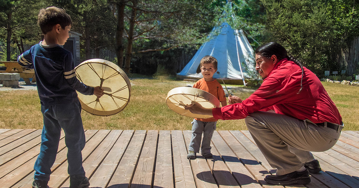 An Ojibwe man teaches two young children about the voice of the drum at the Great Spirit Circle Trail on Manitoulin Island. The Great Spirit Circle Trail is one of the best things to do on Manitoulin Island