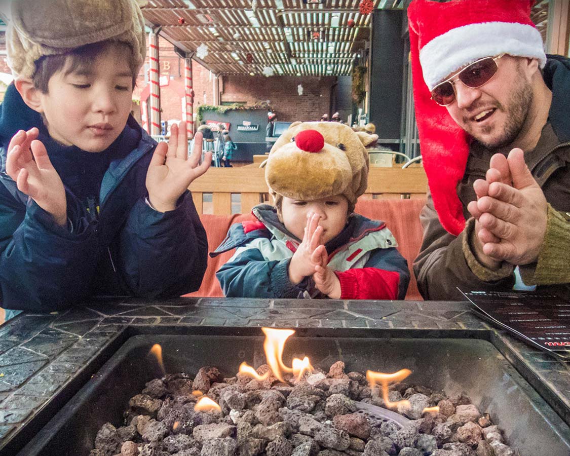 A man wearing a Santa hat and two young boys wearing rudolph hats warm themselves by a fire at a Christmas Market