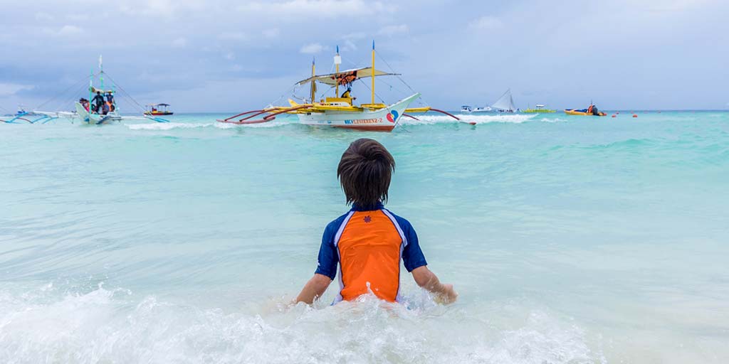 A boy plays in the ocean while Paraw boats float in the distance