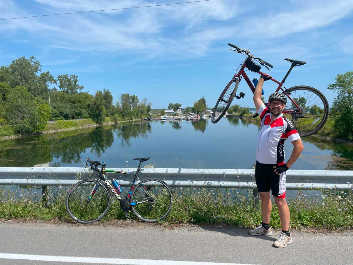 A man in cycling gear lifts a bike over his head along the Cornwall Waterfront Trail