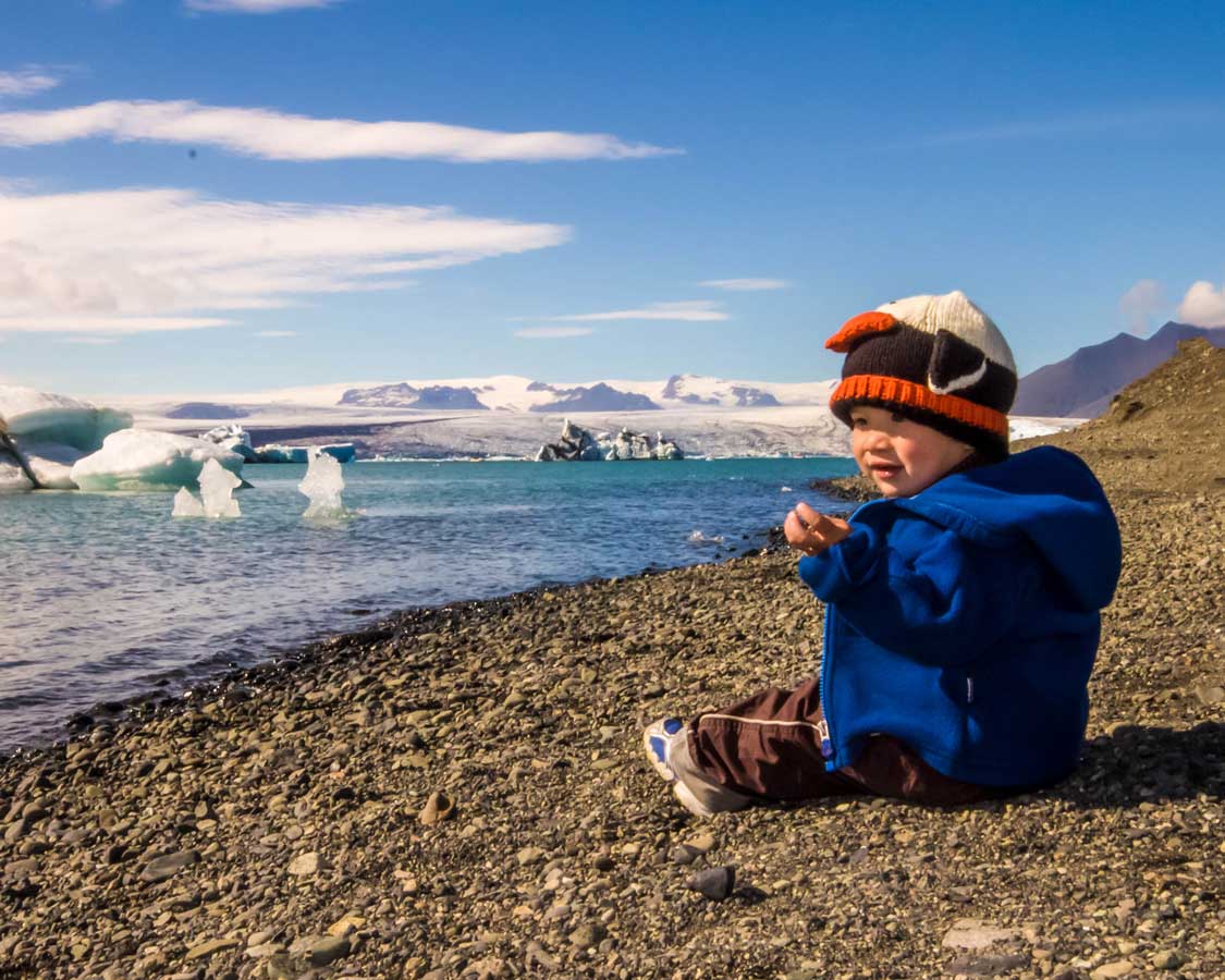 A young boy wearing a penguin hat sits at the edge of a glacier filled lagoon in Iceland for kids