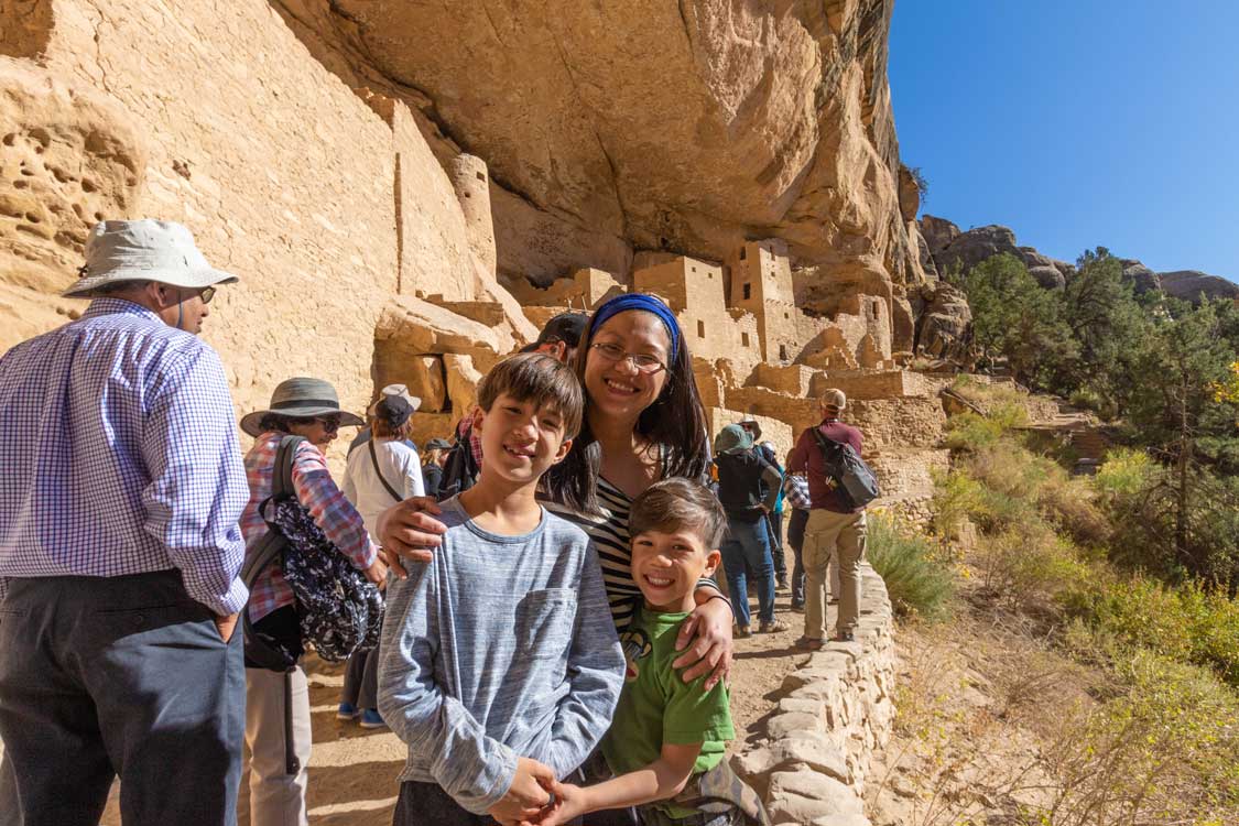 Mother and two sons smile in front of adobe cliffside dwellings in Mesa Verde National Park in Colorado