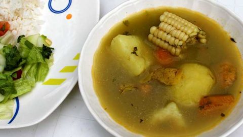 Soup with chicken, potato, and yucca served in a white bowl next to a salad