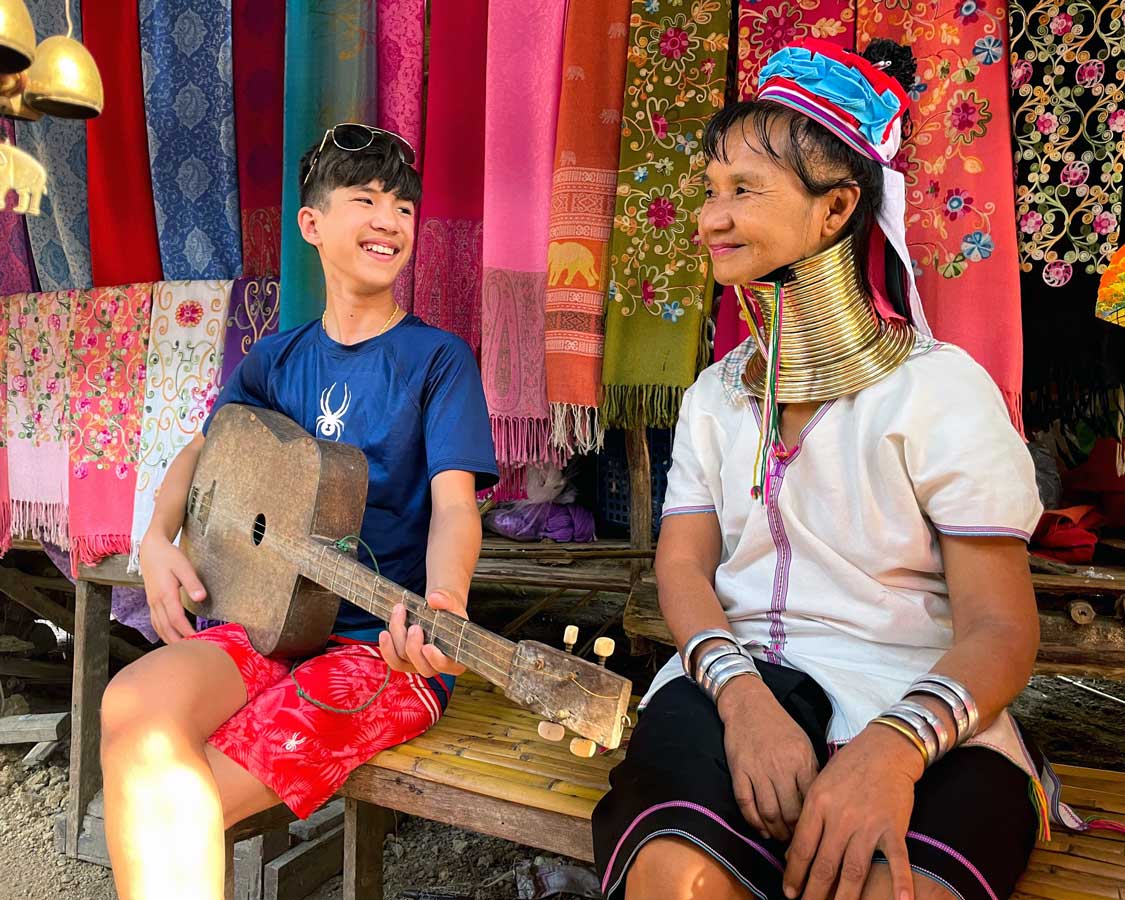 Boy plays guitar with a member of the Long-Necked Karen Hill Tribe in Thailand