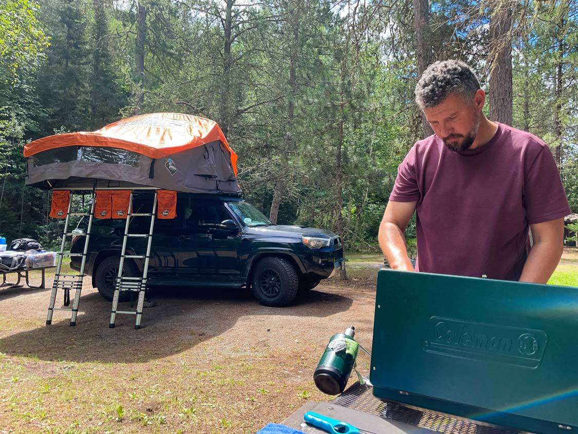 A man cooks on a camp stove in front of a truck with a rooftop tent