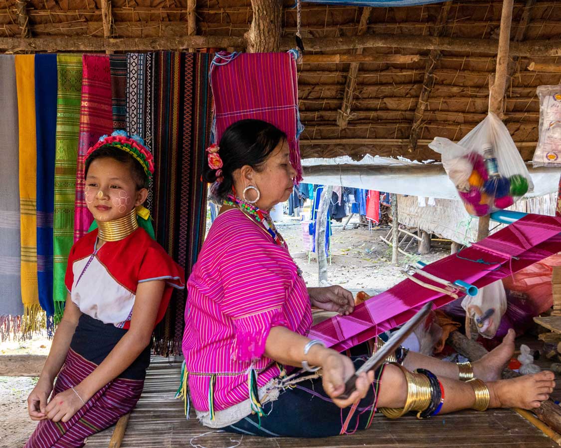 A woman from the White Karen hill tribe weaving next her daughter who is wearing long neck Karen clothes