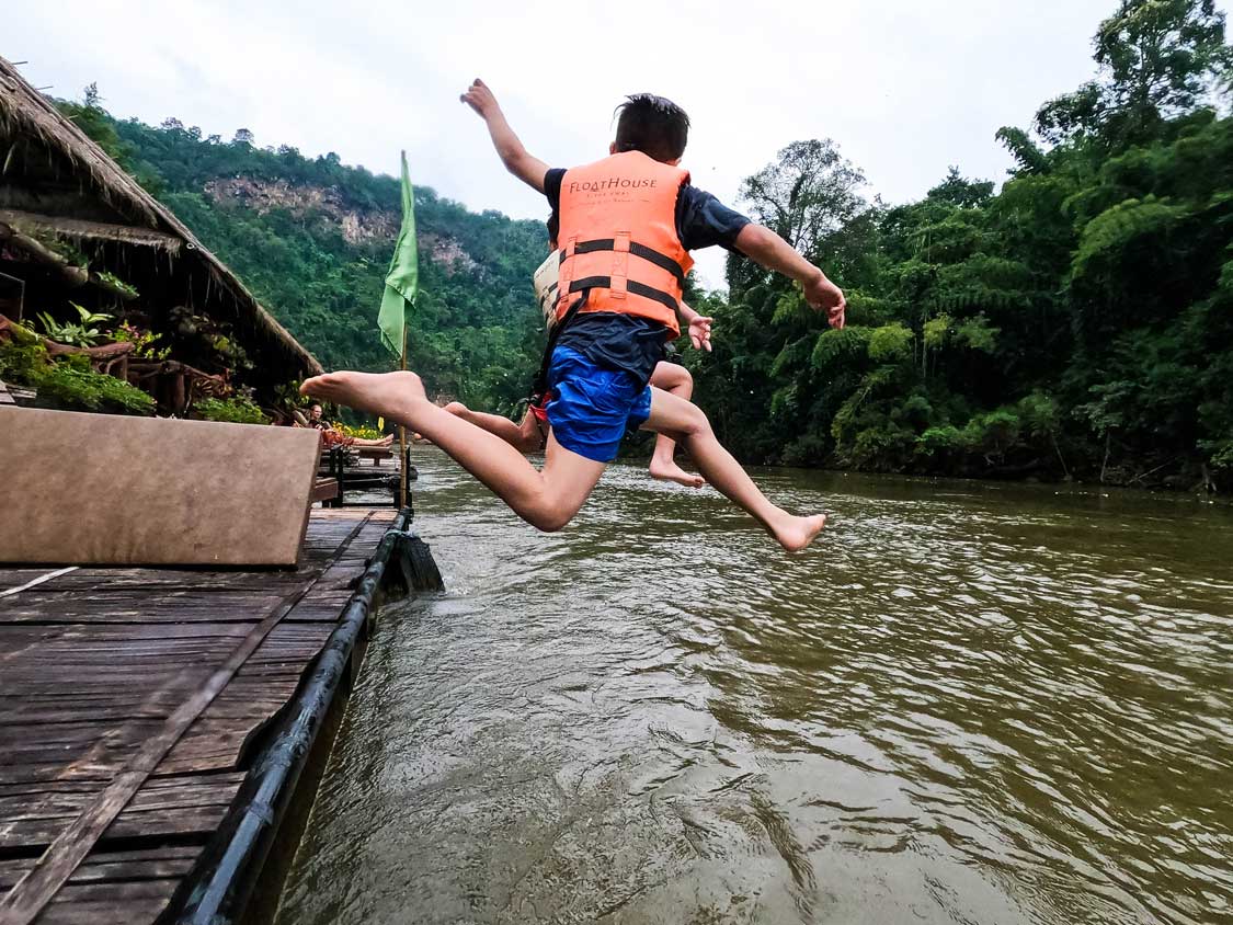 A boy wearing a lifejacket jumps off of a dock into the River Kwai