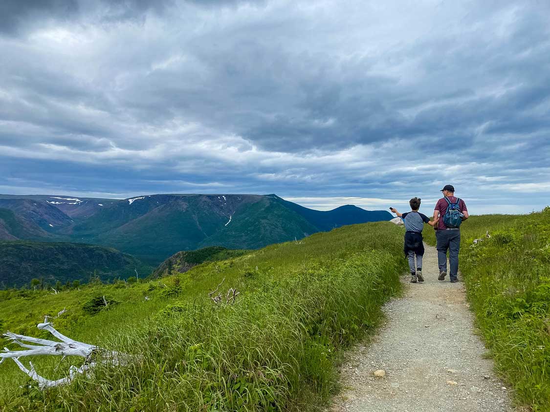 A father and son walk on a hiking trail surrounded by mountains and grass in Gaspesie National Park