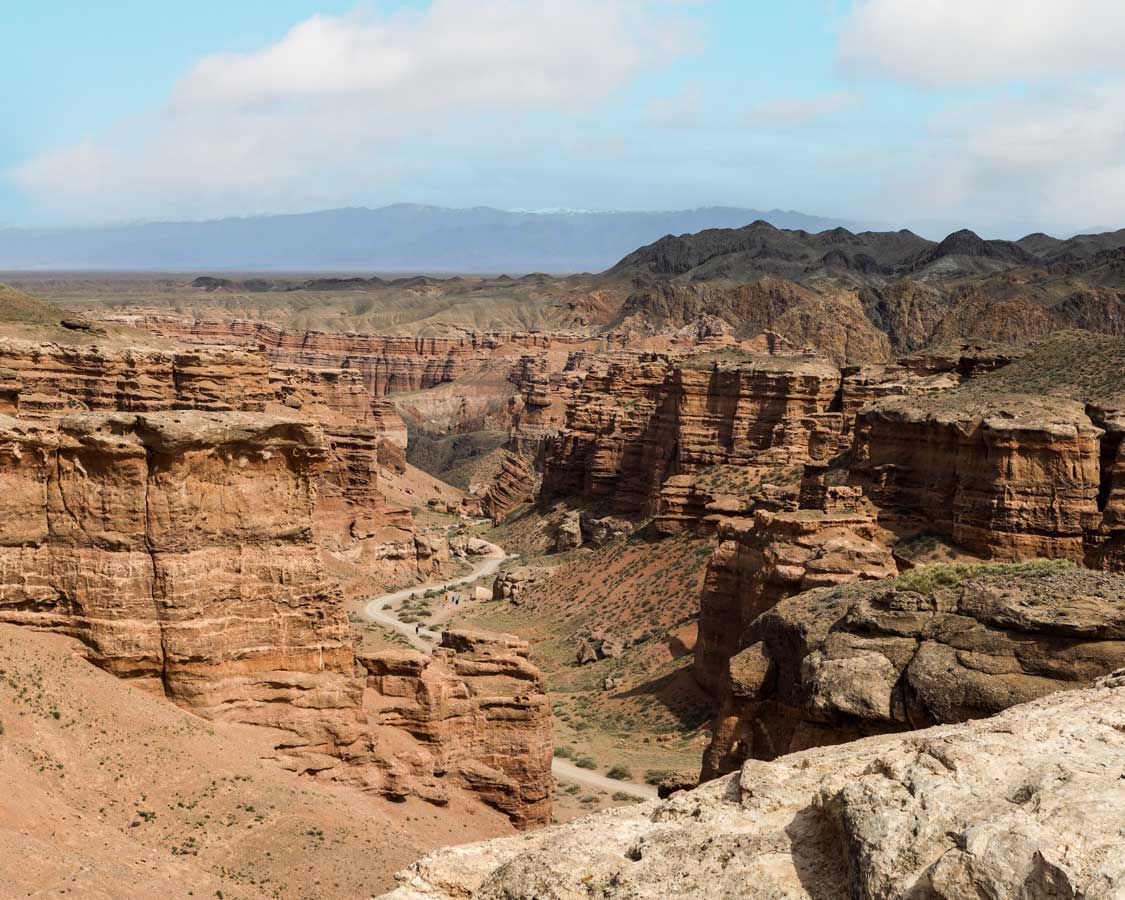 A deep red canyon with dramatic rock formations and a trail at the base where people can be seen walking at Charyn Canyon, Kazakhstan