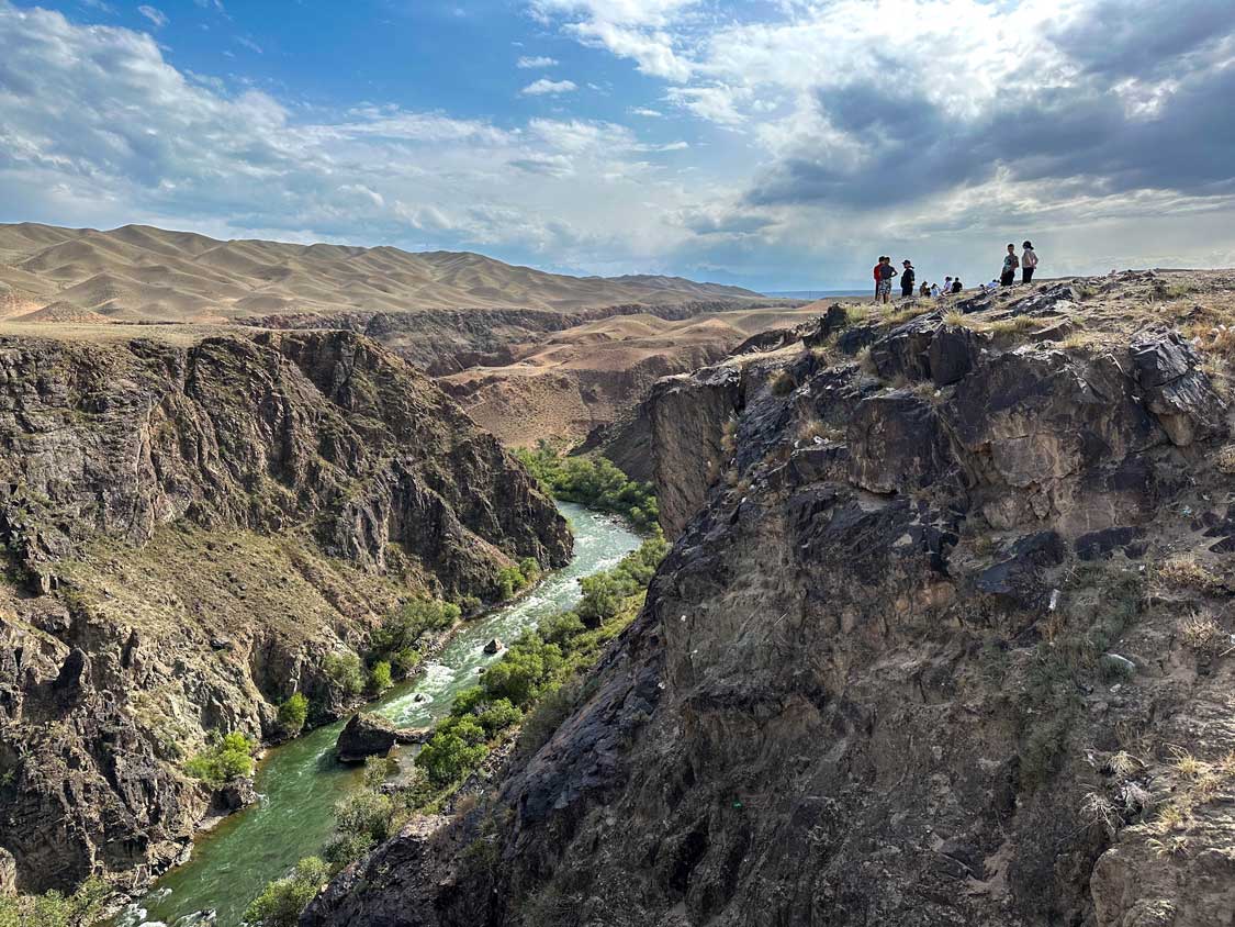 A small crowd of people gaze over the wall of Black Canyon, Kazakhstan to a bright blue river at its base