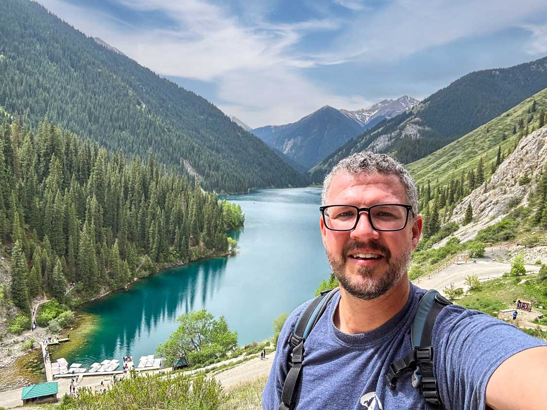 A man takes a selfie in front of a bright-blue mountain lake at Kolsay Lakes National Park