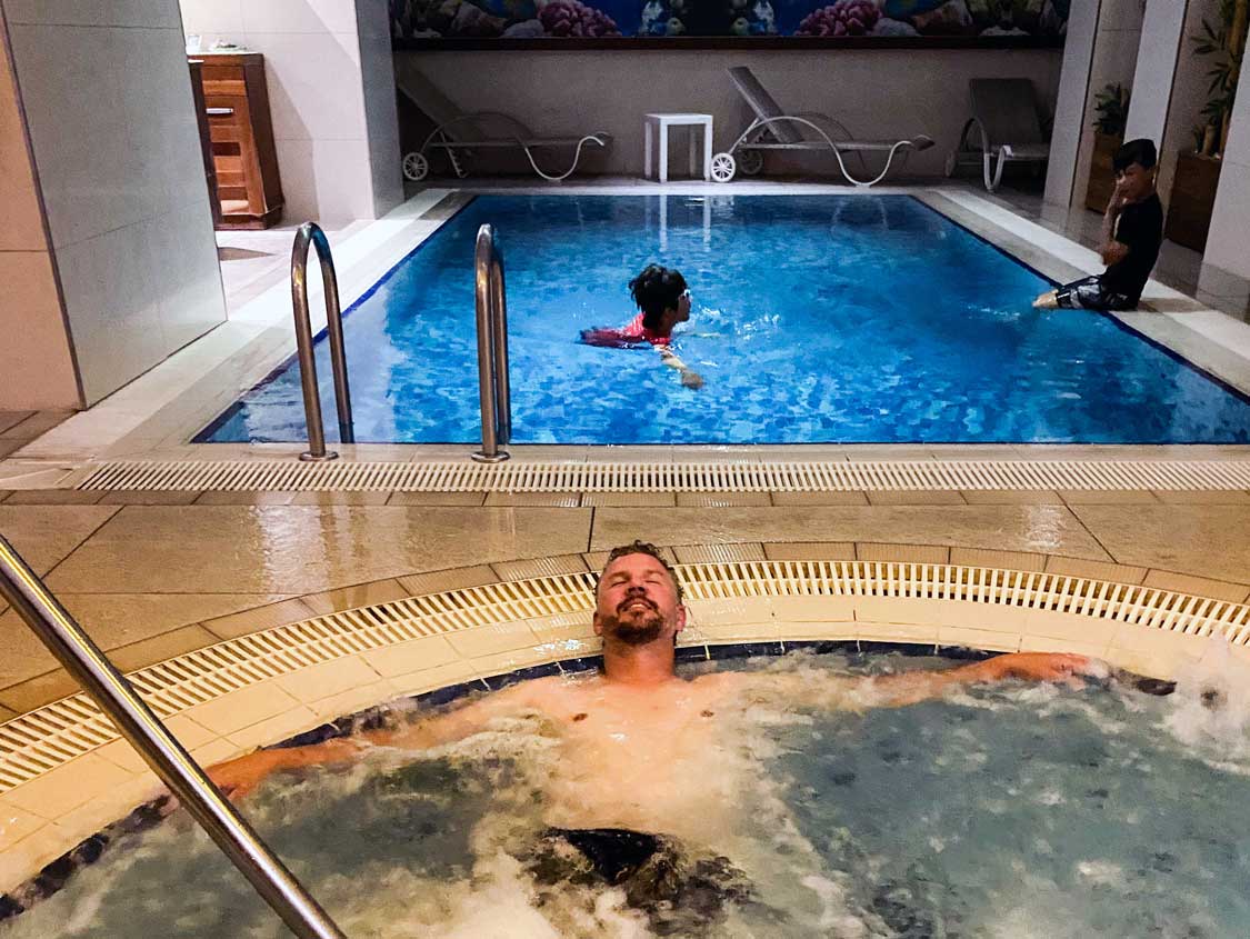 A man relaxes in a hot tub while his children swim behind him