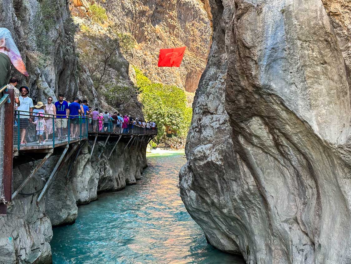 Crowds walk along a cliffside boardwalk above a mountain stream at Saklikent Gorge