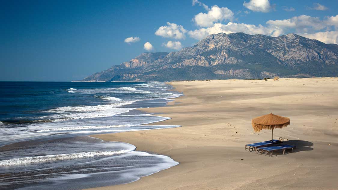 A sun bed with a beach umbrella sit on the sand of Patara Beach, Turkiye with mountains in the background