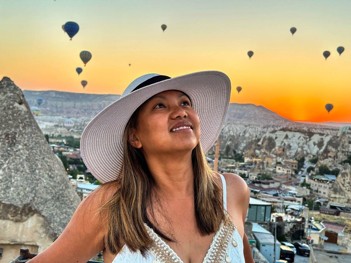 A woman in a white hat looks up at hot air balloons at sunrise in Cappadocia, Turkiye