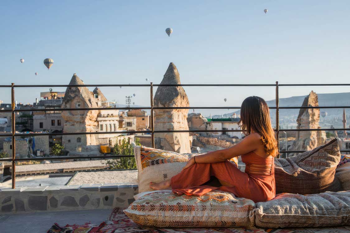 A woman relaxes on the terrace of the Lunar Cappadocia cave hotel while hot air balloons fill the sky