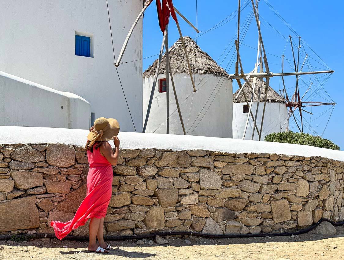 A woman in a red dress holds a floppy hat in front of the Mykonos windmills