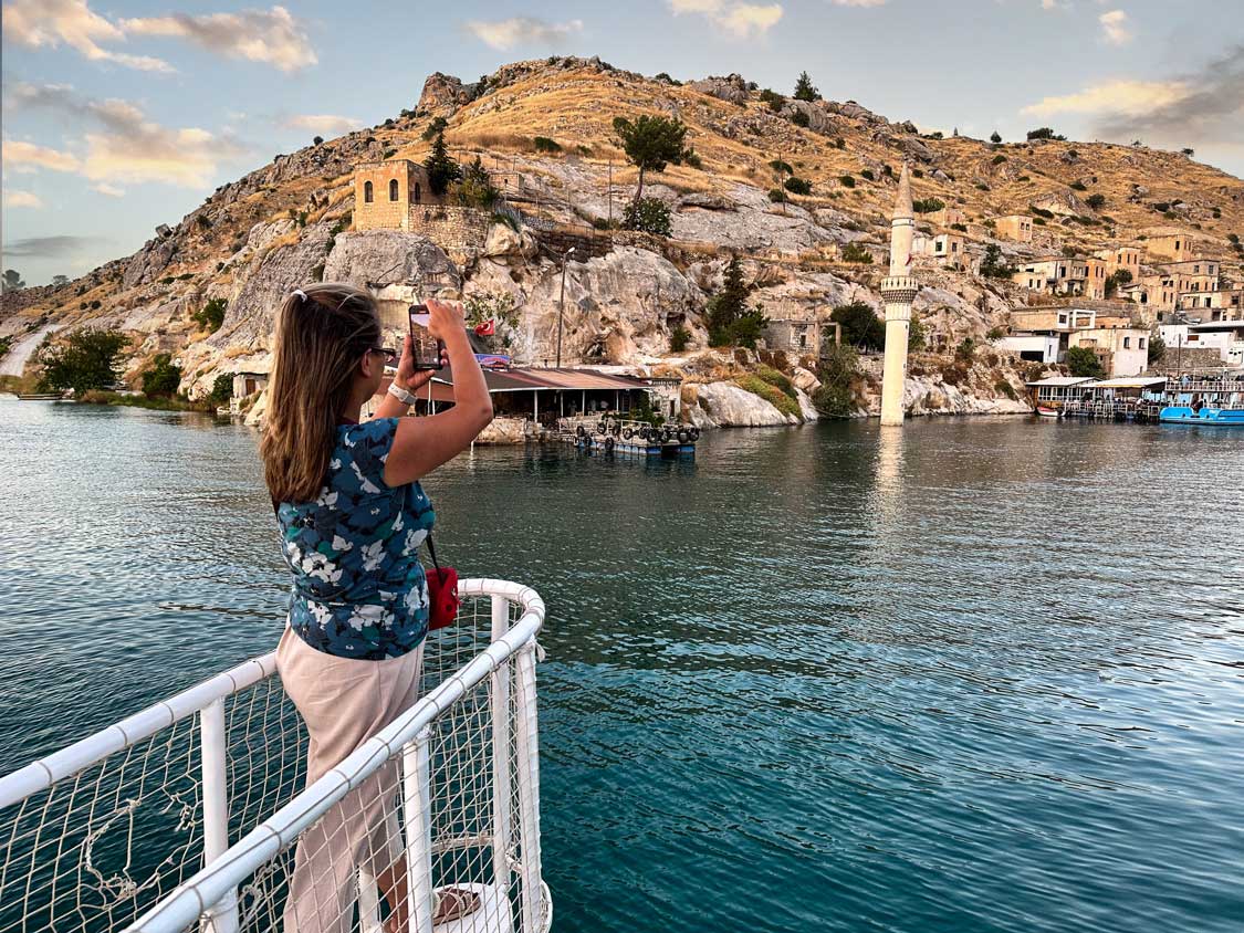 A woman takes a photo of the Halfeti Sunket City from the front of a boat.