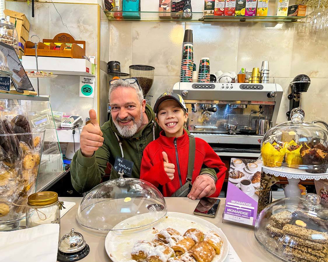 A boy smiles behind the counter in the arms of Thanos, the owner of Migniardise Cafe in Athens, Greece