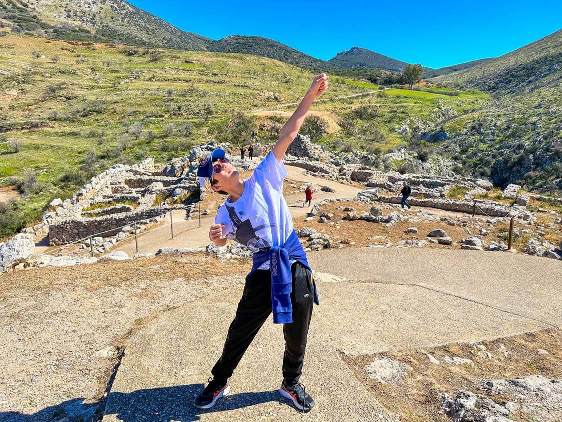 A preteen boy makes a dramatic pose while exploring the Mycenae ruins in Greece.