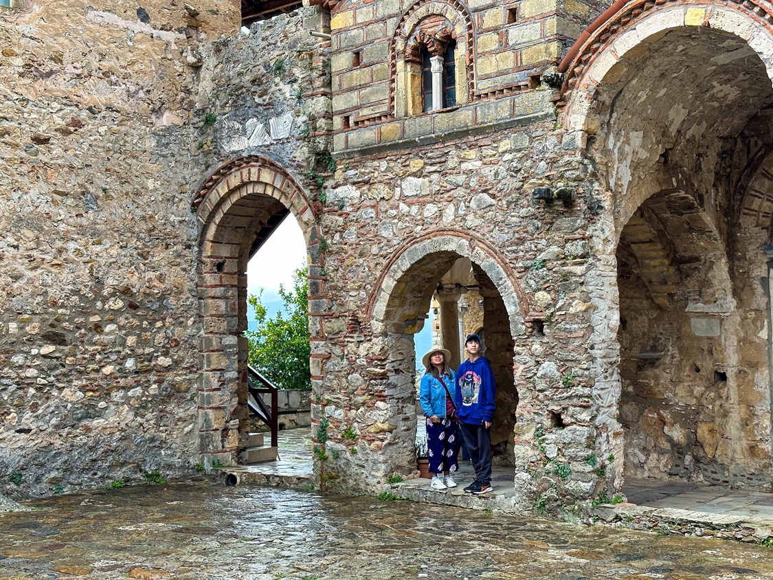 A mother and son look out from a crumbling church tower in Mystras Greece at a rainy courtyard