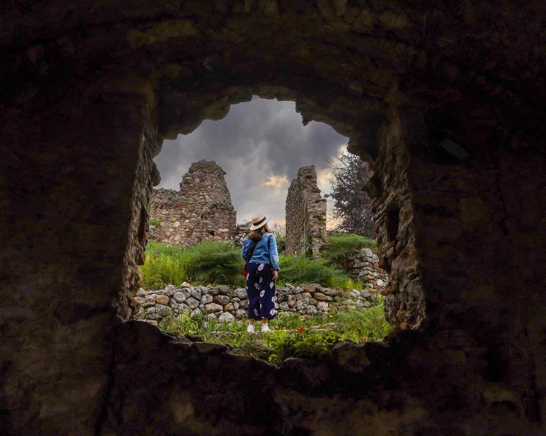 A woman framed by a ruined wall looks up at a cloudy sky in Mystras, Greece