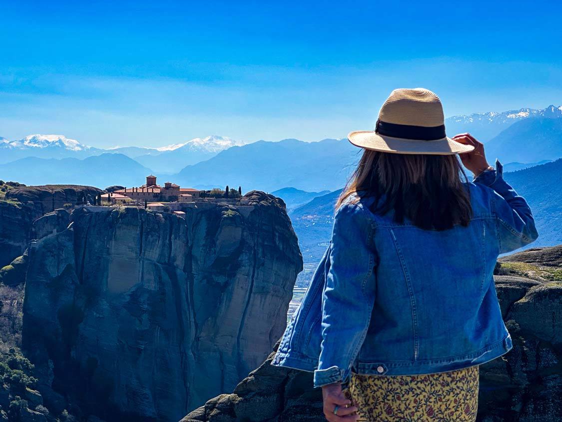 A woman holds her hat while looking across a gorge at the Holy Monastery of Saint Stephen in Meteora, Greece