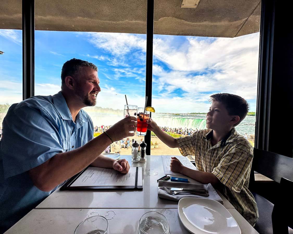 A father and son cheers at Table Rock Restaurant with Niagara Falls in the background