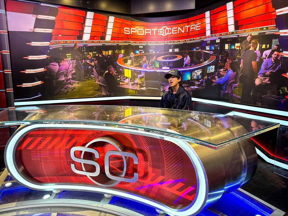 A boy in a hockey jacket sits behind the desk of the SportsCentre Broadcast Booth a the Hockey Hall of Fame