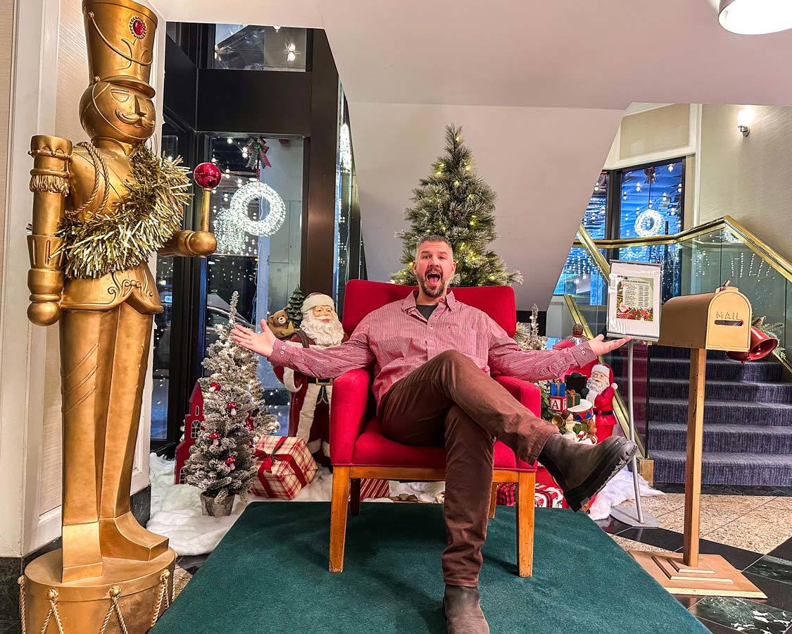 A man in a red shirt smiles at the camera while sitting in a Santa display at the Chelsea Hotel in Toronto