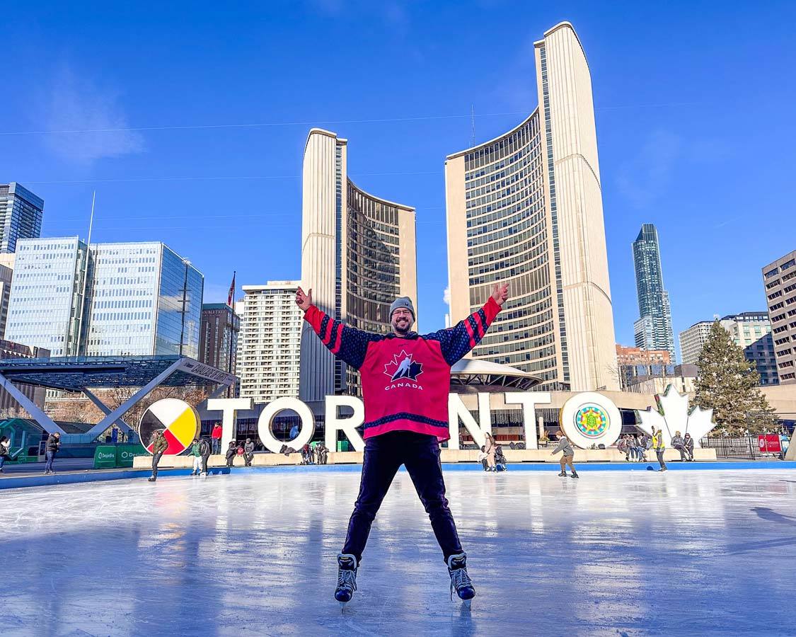 A man wearing a Team Canada Jersey smiles while skating at Nathan Phillips Square during winter in Toronto