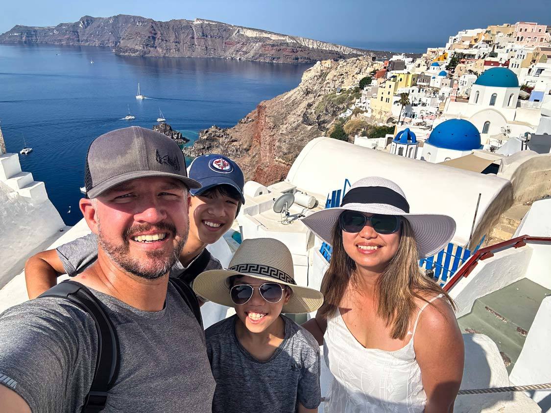 The Wandering Wagars Family smiles from a lookout over the blue roofed buildings in Santorini, Greece