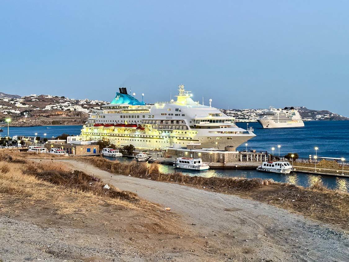 A Celestyal cruise ship is docked at the port of Mykonos at sunrise with the massive Odyssey of the Seas in the background