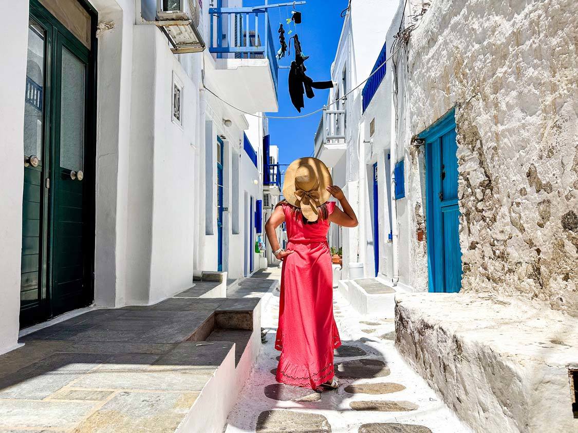 A woman wearing a pink dress and straw hat looks up at fresh laundry hanging over an alley while walking the streets of Chora, Mykonos